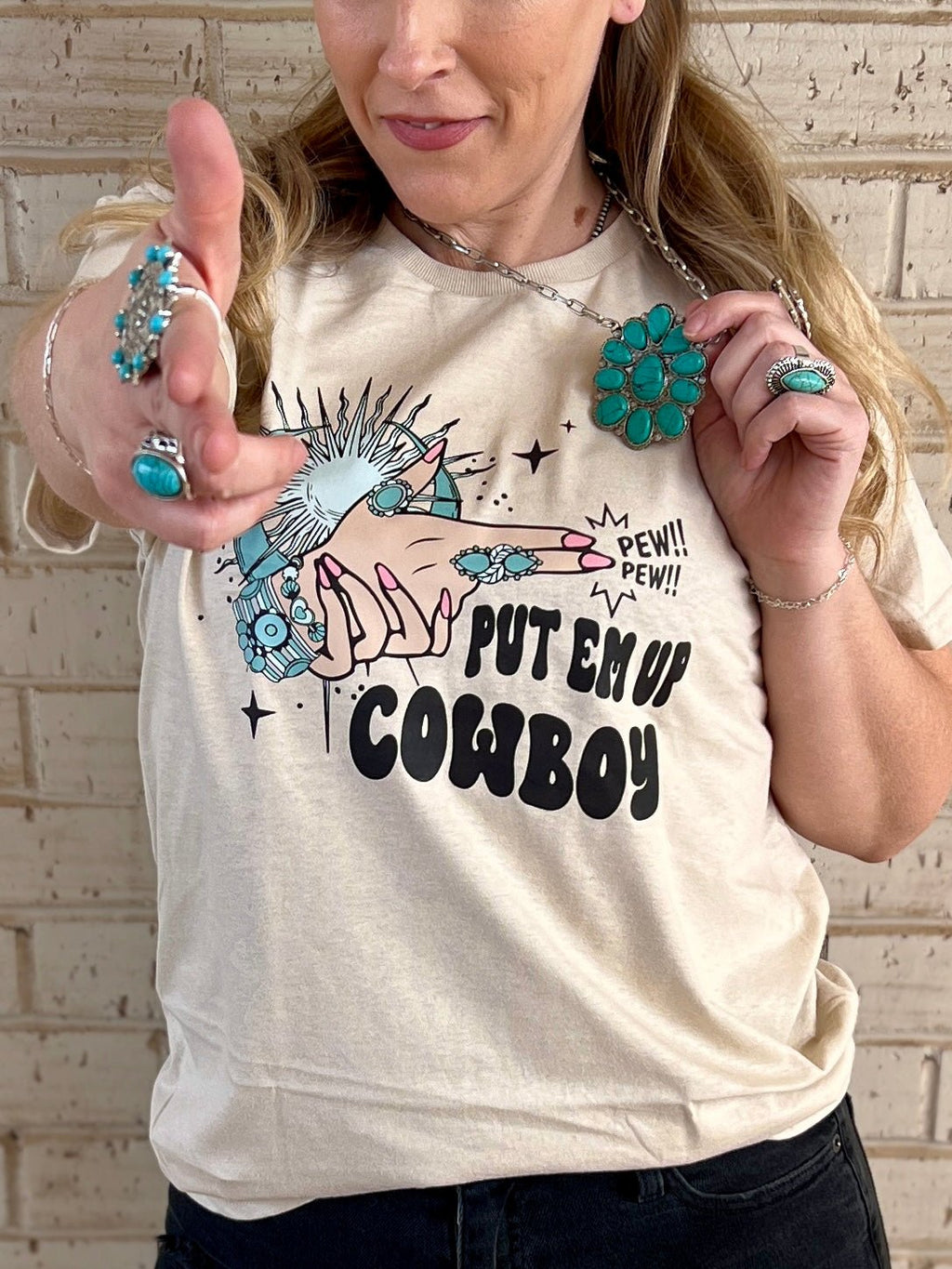 Western style. Western graphic tees. Western t-shirts. "Put em' up cowboy" shirt. Western t-shirts. Shirt with a skeleton hand on it. Shirts with turquoise rings on it. Western shirts. Graphic T-shirts. Graphic tees. Western boutique. Women's western graphic tees. Women's western boutique. Small business. Woman owned.