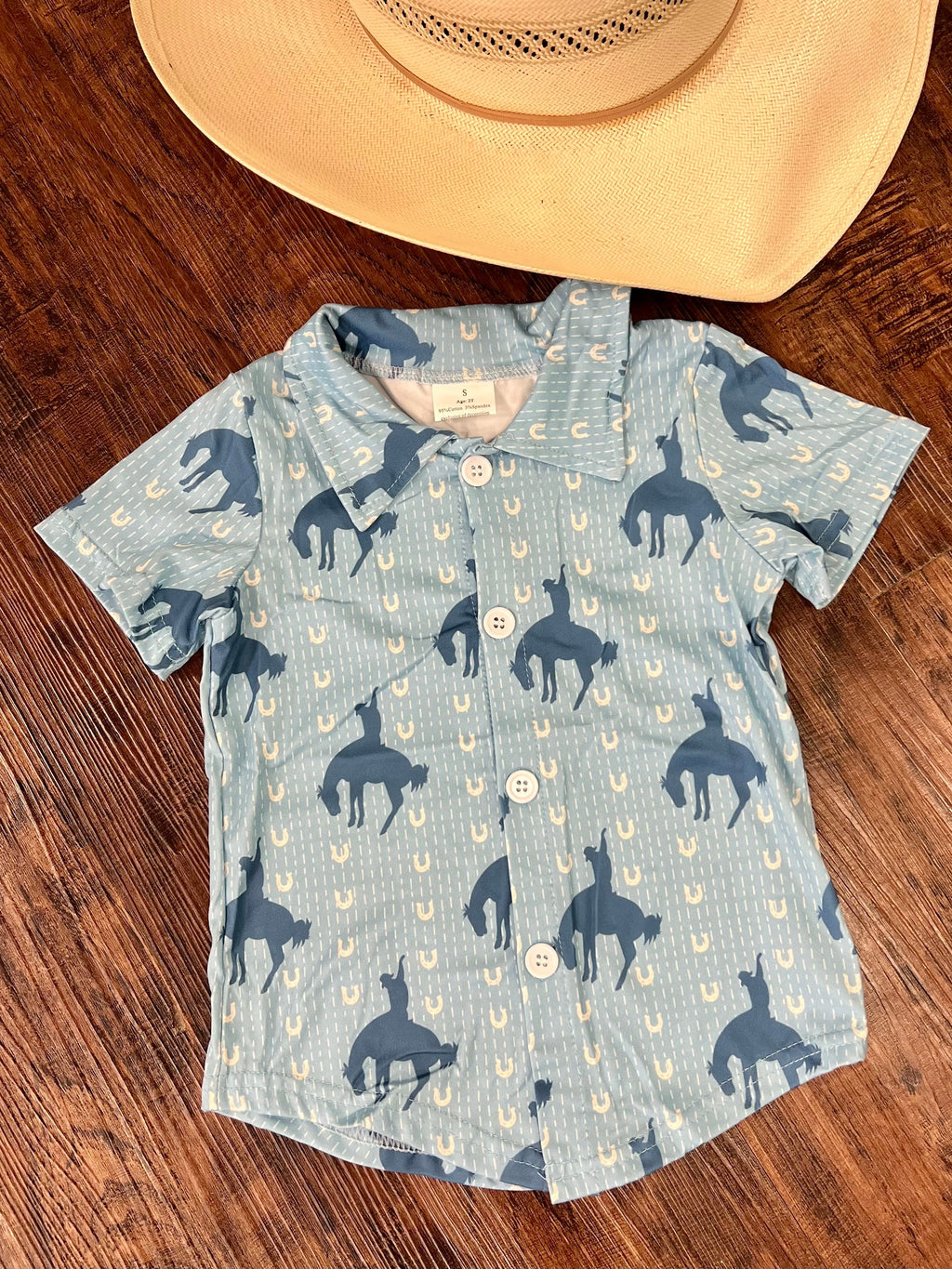 This Kids Lucky Cowboy Button Up is the perfect way to add a dash of western style to your little one's wardrobe! In a baby blue hue, this short-sleeved collared shirt will have them galloping around in style with its bucking-horse, horseshoe detailing. Yeehaw!  95% Cotton, 5% Spandex