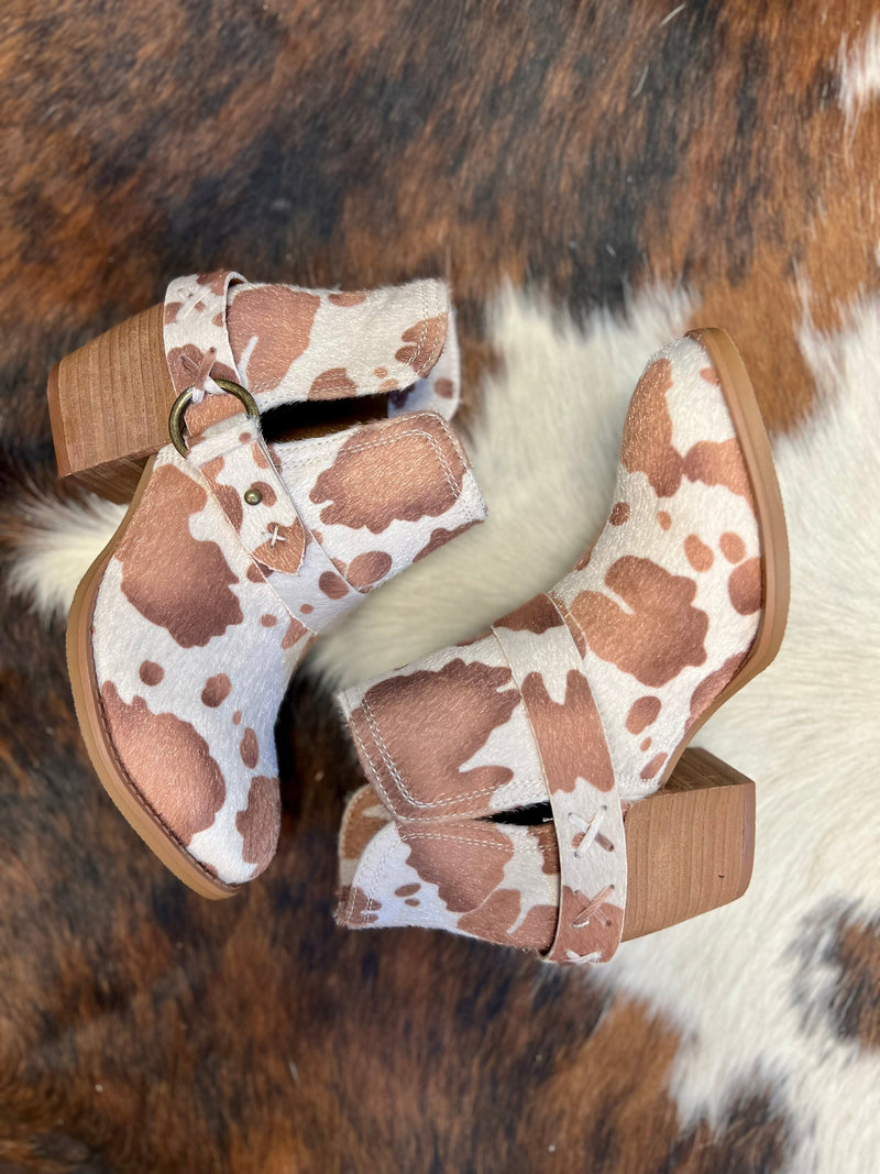 Step up your style game with the Moove Out The Pasture Booties! Featuring a bold  light brown and white cow print faux fur, 2 1/2" wood heel, 6" total height, plus inside and outside ankle slits and a wrap around ankle strap, these booties will take your look to the next level. Show ‘em you mean business!