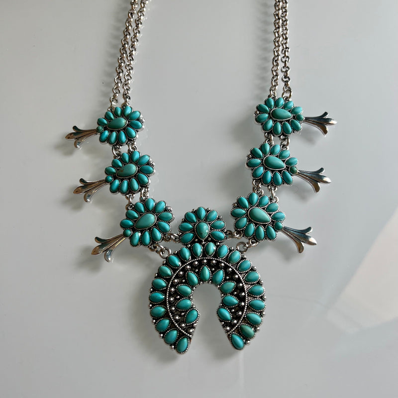This statement piece is sure to make a statement at any event: the It Takes Two Squash Blossom Necklace. Features include a choice of turquoise or turquoise and white, a 22" silver chain with a 3" adjustable clasp, 7 mini stone conchos for added character, and a double chain with a 2" squash blossom. Feel fashionable and distinctive!