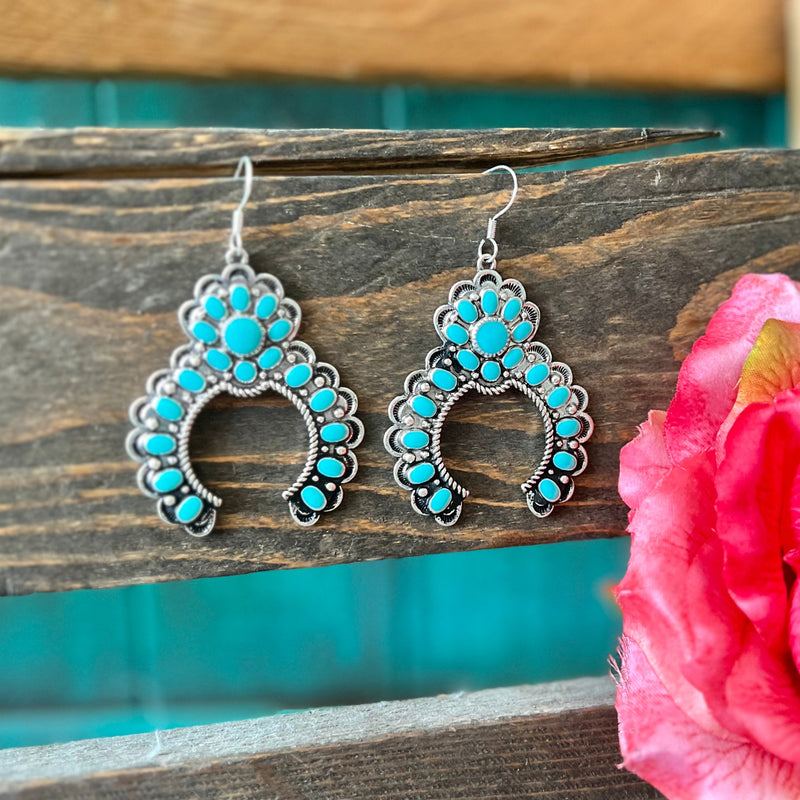 These In Full Bloom Blossom Earrings are crafted from high polish silver in a western concho style. The textured squash blossom design features genuine turquoise stones and a fish hook dangle for a unique, eye-catching look. Perfect for any occasion.