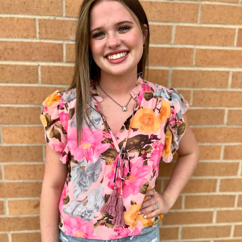 This Floral Vanity Blouse is perfect for summer days. With its pink floral print, tassel tie, and short sleeve cut, it's sure to make a statement. It has ruffle detailing around the neck and a notched V neckline for an elevated look. Made from 100% Polyester, it's lightweight and breathable.