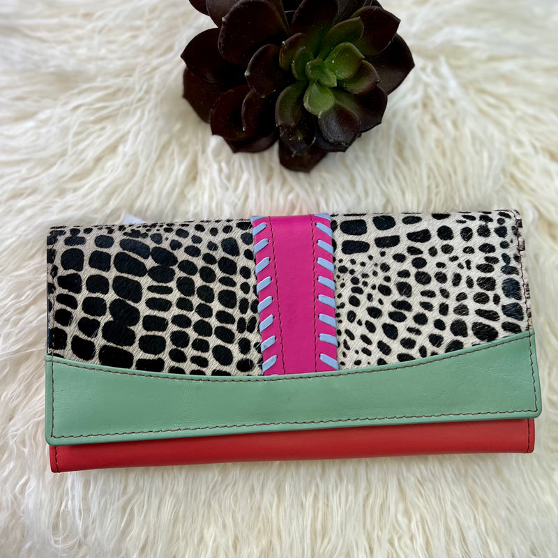 Step out in style with this one-of-a-kind Brooks Leather Wallet. Crafted with exquisite care from black and white spotted hide, this eye-catching wallet features a chic bright pink leather detail offset by a purple buckstitch. It's further adorned with blue and red leather which adds a note of sophistication to the piece. Ideal for stashing your cards and currency safely, it offers a fold over flap with snap closure for security, and measures 8"W x 4"H. An ideal accessory for the modern trendsetter.