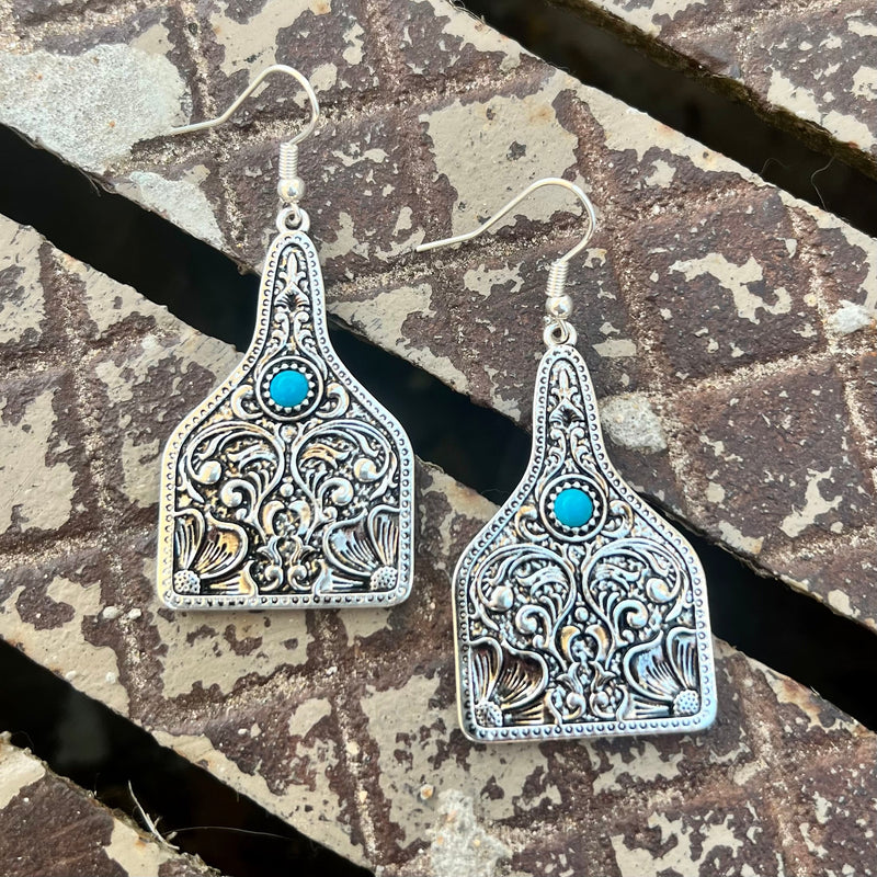 Add a luxurious touch to your look with our Paisley & Turquoise Tags. Handcrafted with a delicate silver paisley scroll design, these tags feature a classic cattle tag and dangle from a beautiful fish hook with a stunning turquoise stone. With a length of 2", these tags make the perfect statement piece.