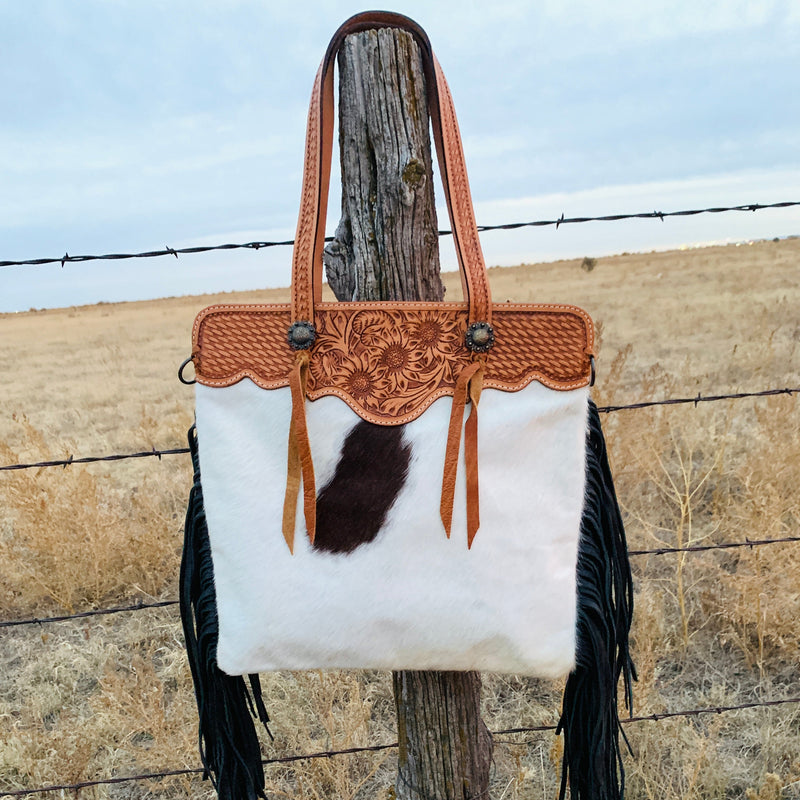 Hair on hide leather shoulder bag with hand tooled straps and top trim. Features decorative concho with tassel and black suede fringe down sides. One large outside zipper pocket, one large inside zipper pocket, and two inner open pockets. Back is black leather. Includes removable cross body strap.  H17"xW17"  10" handles