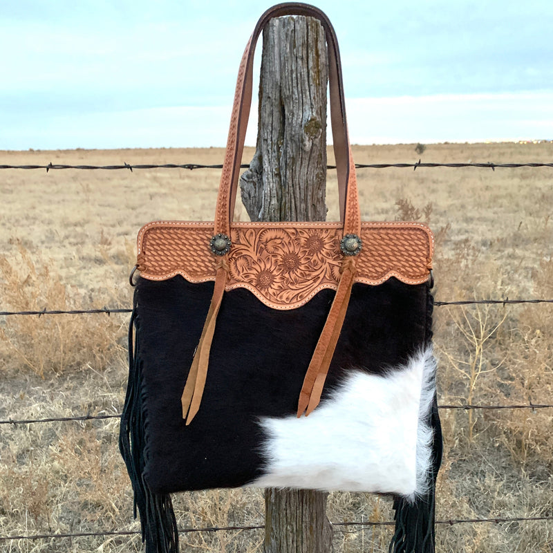 Hair on hide leather shoulder bag with hand tooled straps and top trim. Features decorative concho with tassel and black suede fringe down sides. One large outside zipper pocket, one large inside zipper pocket, and two inner open pockets. Back is black leather. Includes removable cross body strap.  H17"xW17"  10" handles