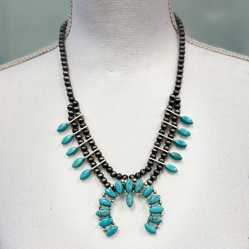 The "Don Quijote" Necklace is a 2.5" Wide 14 bead turquoise pendant. There are 5 turquoise pear stones detailed on the sides attached to the Buffalo Pearl Strand Necklace. The buffalo pearl strand necklace is a 10" strand with adjustable clasp.