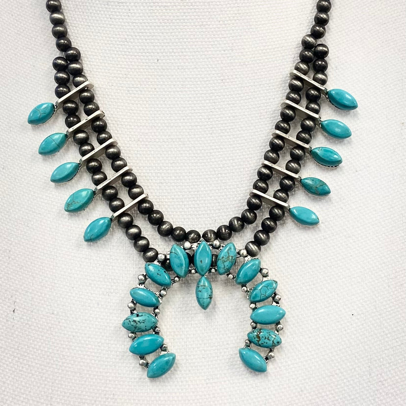 The "Don Quijote" Necklace is a 2.5" Wide 14 bead turquoise pendant. There are 5 turquoise pear stones detailed on the sides attached to the Buffalo Pearl Strand Necklace. The buffalo pearl strand necklace is a 10" strand with adjustable clasp.