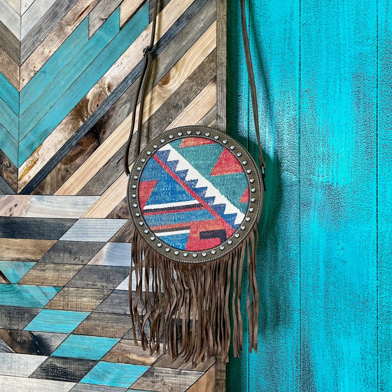 The Paradise Valley Canteen Bag is a 10" round multi colored Aztec Print Bag. The bag features a 3/4 zipper closure and a small zipper closure on the back. The bag has brown leather fringe and silver nail head detail.   10" round in size  48" adjustable strap 
