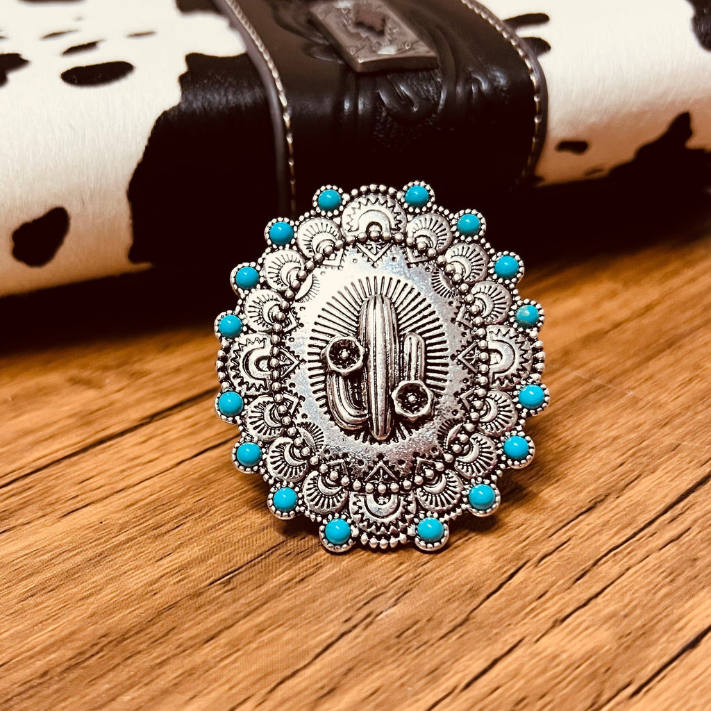 The Big Cactus Stretch Ring is a 2" round western stamped ring with a high polish textured western style concho Cactus placed at it's center. The ring features a western stamped design with small turquoise stones placed around the edge.  Flexible and adjustable ring cuff.   2" pendent 