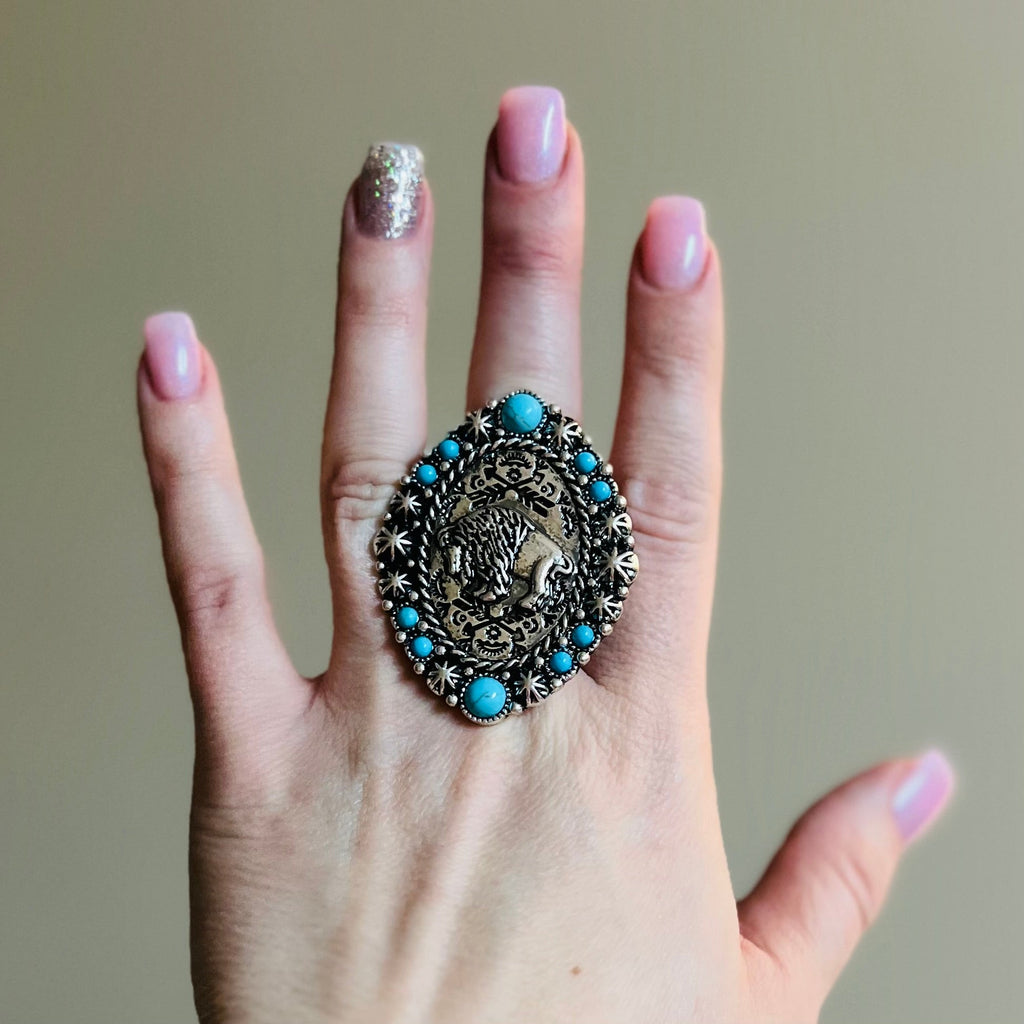 The Big Bison Stretch Ring is a 2" round western stamped ring with a high polish textured western style concho Bison placed at it's center. The ring features a western stamped design with small turquoise stones placed around the edge.  Flexible and adjustable ring cuff.   2" pendent 