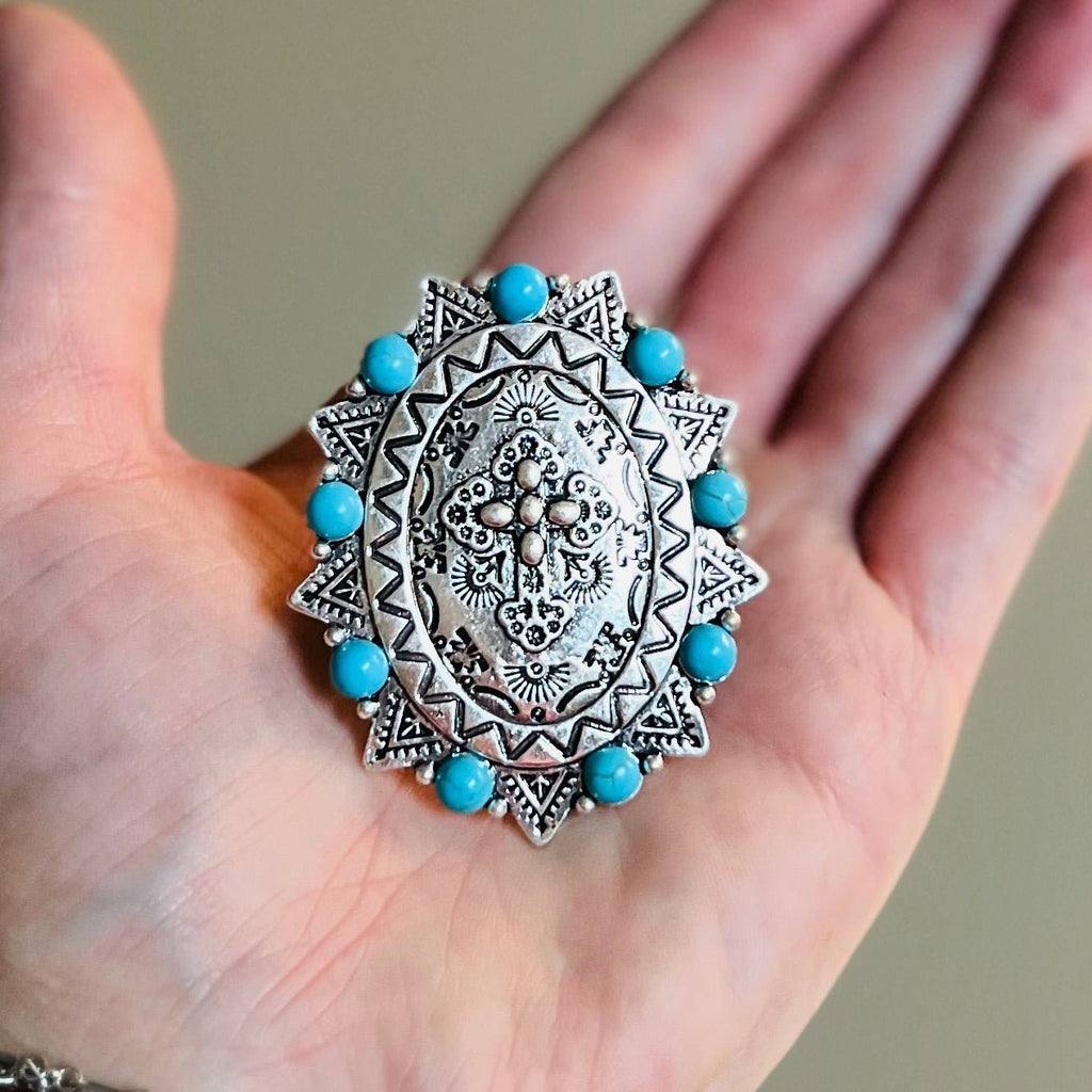 The Big Cross Stretch Ring is a 2" round western stamped ring with a high polish textured western style concho Cross placed at it's center. The ring features a western stamped design with small turquoise stones placed around the edge.  Flexible and adjustable ring cuff.   2" pendent 