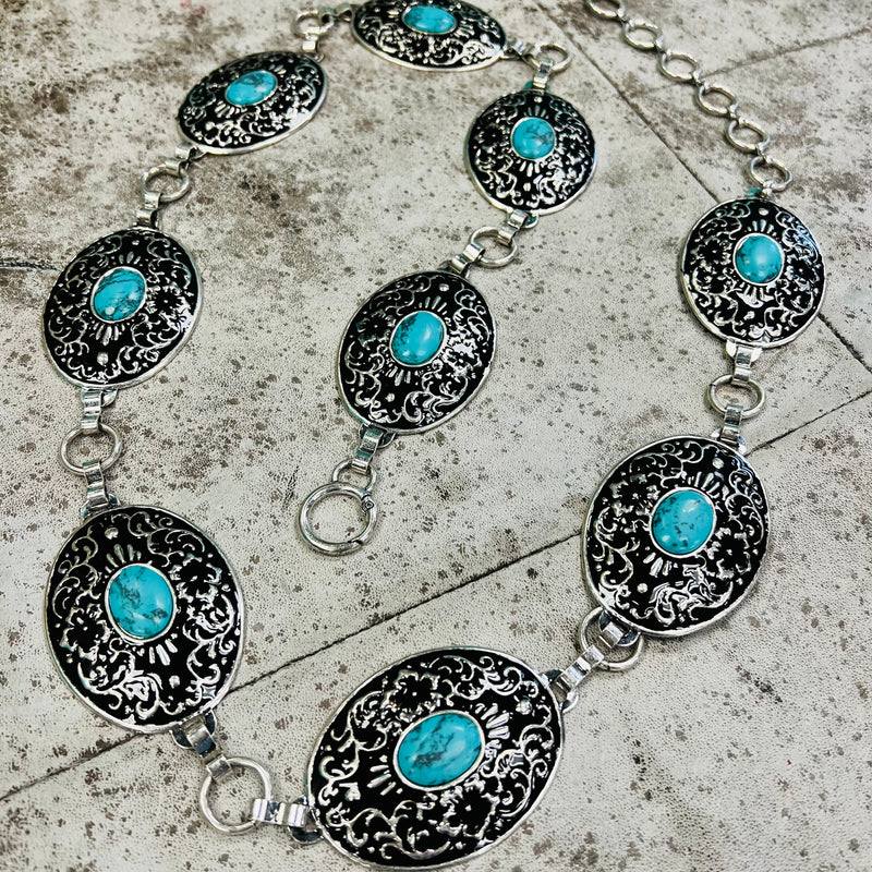 The At the Mountain Top Chain Link Belt is a silver and turquoise stone concho belt. The concho's have a black background with a floral scroll pattern design. The concho's are 2" X 2 1/2" in size. And the belt is 46" long with the adjustable chain links. 