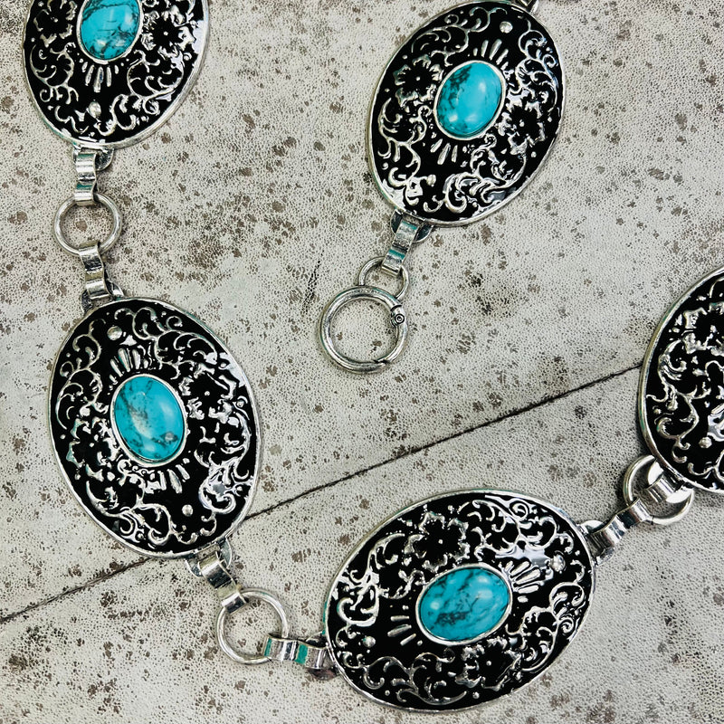 The At the Mountain Top Chain Link Belt is a silver and turquoise stone concho belt. The concho's have a black background with a floral scroll pattern design. The concho's are 2" X 2 1/2" in size. And the belt is 46" long with the adjustable chain links. 