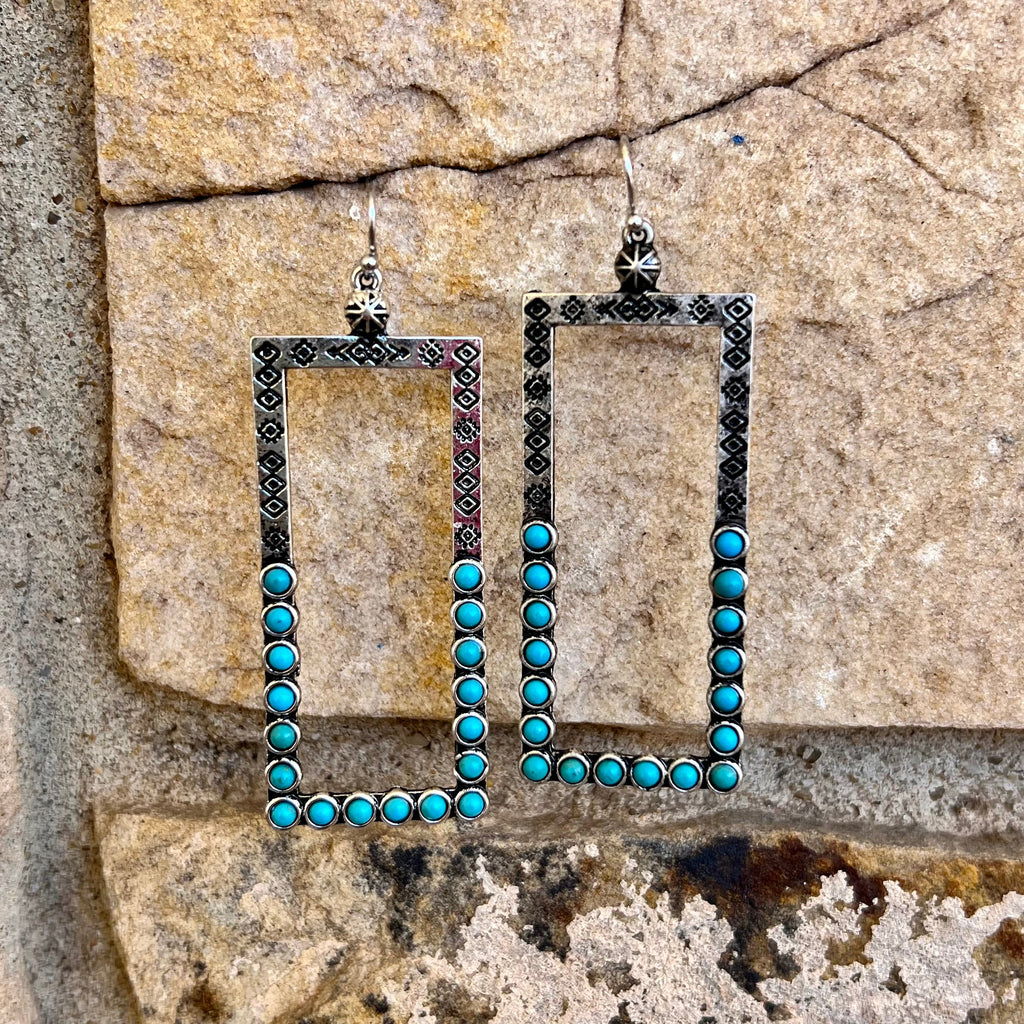 These rustic western In The Chute Earrings feature a high polish square bar casting and genuine turquoise stones. With their eye-catching design, these stylish earrings are the perfect way to add a touch of southwestern flair to any outfit.  2 1/2" in length