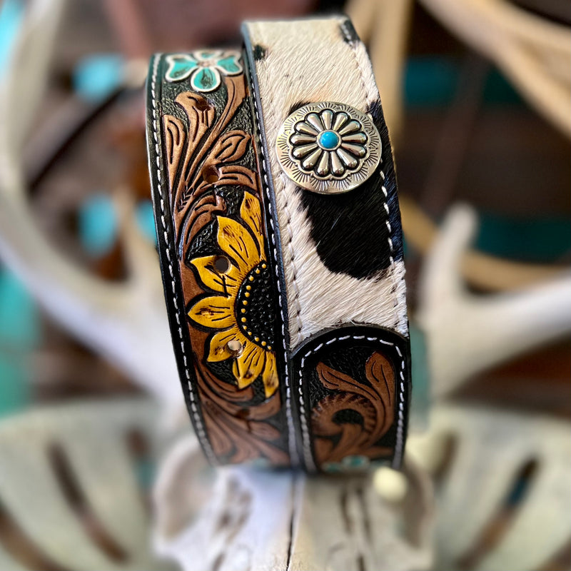 This Bareback in a Sunflower Field Belt features black and white hair on hide detail that's been expertly tooled and painted for a unique look. The belt is finished with a black leather strap, silver flower concho with turquoise stone, and a square belt buckle with turquoise stone. An eye-catching statement piece!  Small-35", Medium-37", Large-39", XLarge-41"