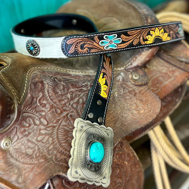 This Bareback in a Sunflower Field Belt features black and white hair on hide detail that's been expertly tooled and painted for a unique look. The belt is finished with a black leather strap, silver flower concho with turquoise stone, and a square belt buckle with turquoise stone. An eye-catching statement piece!  Small-35", Medium-37", Large-39", XLarge-41"