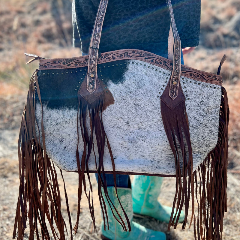 A western bag as unique as the horse you rode in on! Oversized hair on hide satchel bag that is accented with oiled tooled leather surrounded in small gold studs - plus leather fringe and tassels! The concealed carry pocket on the back is just the cherry on top! Brown leather back and bottom. Rounded top with zipper closure.  19"Wx13"Hx6"D  handle: 8"