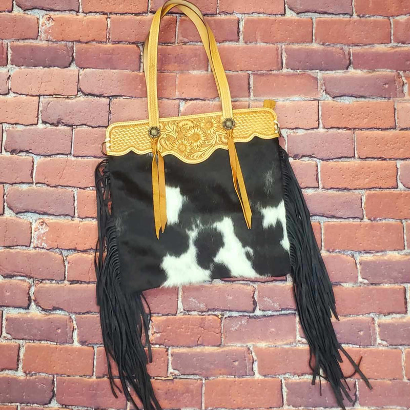 Hair on hide leather shoulder bag with hand tooled straps and top trim. Features decorative concho with tassel and black suede fringe down sides. One large outside zipper pocket, one large inside zipper pocket, and two inner open pockets. Back is black leather. Includes removable cross body strap. H17"xW17" 10" handles