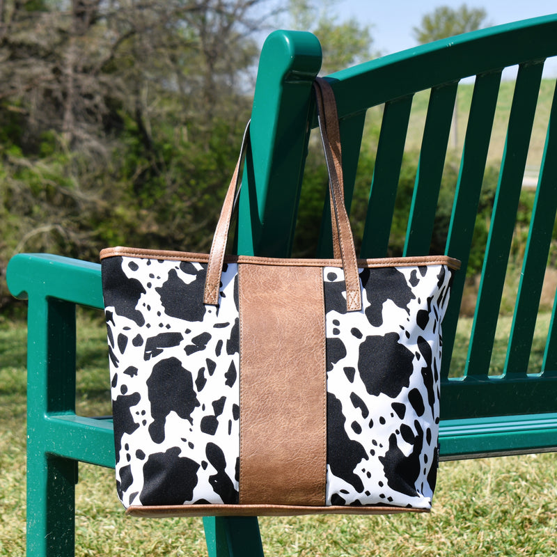 Canvas and vinyl small tote or shopper bag with zipper top closure. Cow print fabric and tan faux leather center strip and handles.