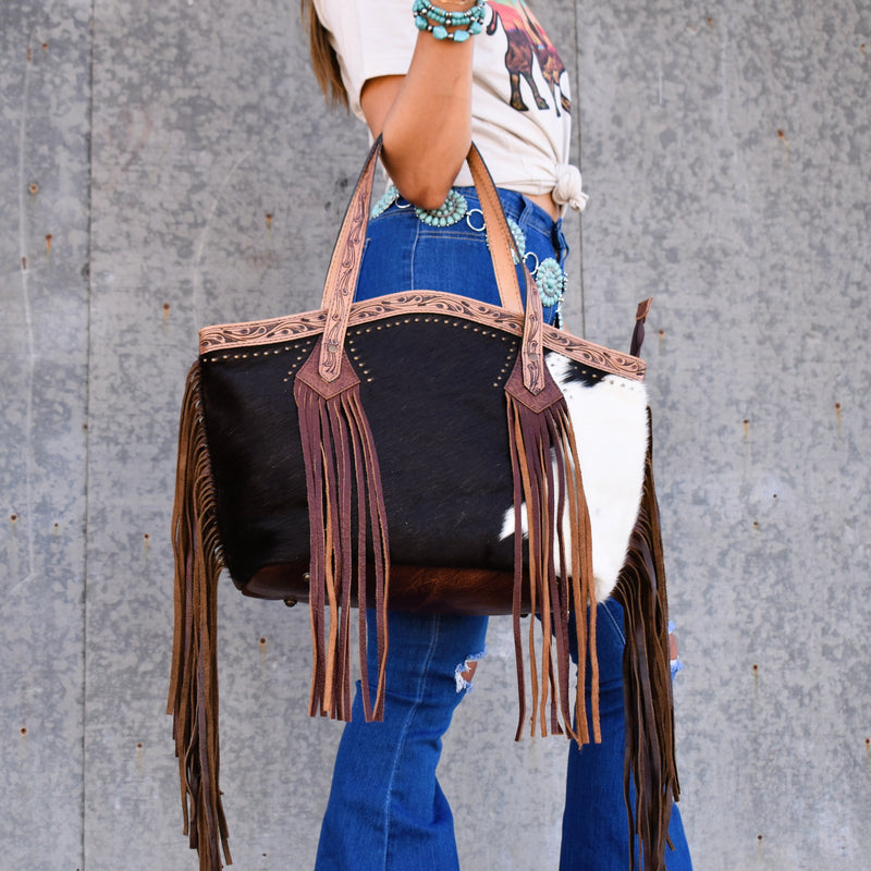 A western bag as unique as the horse you rode in on! Oversized hair on hide satchel bag that is accented with oiled tooled leather surrounded in small gold studs - plus leather fringe and tassels! The concealed carry pocket on the back is just the cherry on top! Brown leather back and bottom. Rounded top with zipper closure.