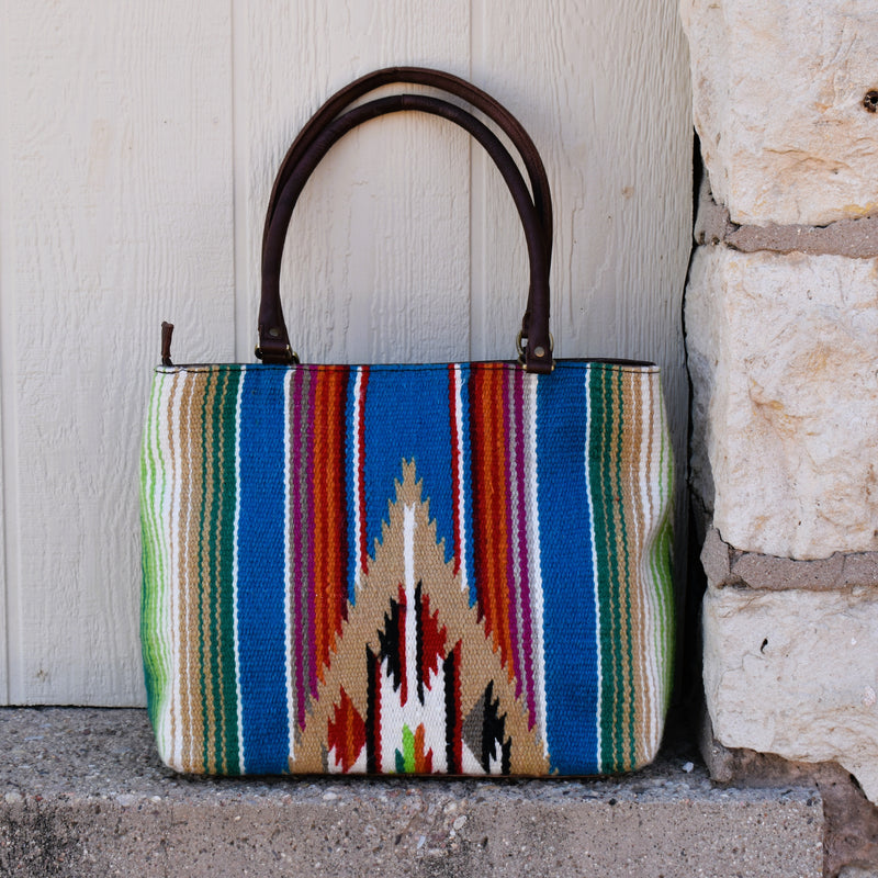 Multicolored serape saddle blanket bag with white buckstitch tooled leather top detail in the front. Padded double handles that are perfect to rest on your shoulder. Brass feet for durability and cleanliness. Zipper top closure.